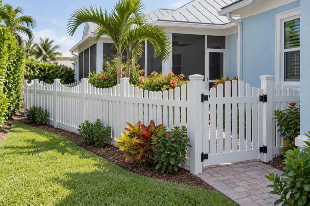Scalloped vinyl pickets and a gate beside a pale blue stucco coastal-style home.
